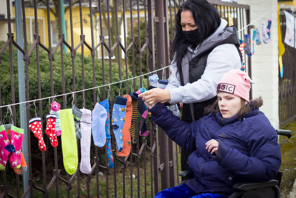 Opiekunka z dzieckiem na wózku inwalidzkim wieszają kolorowe skarpetki na ogrodzeniu szkoły